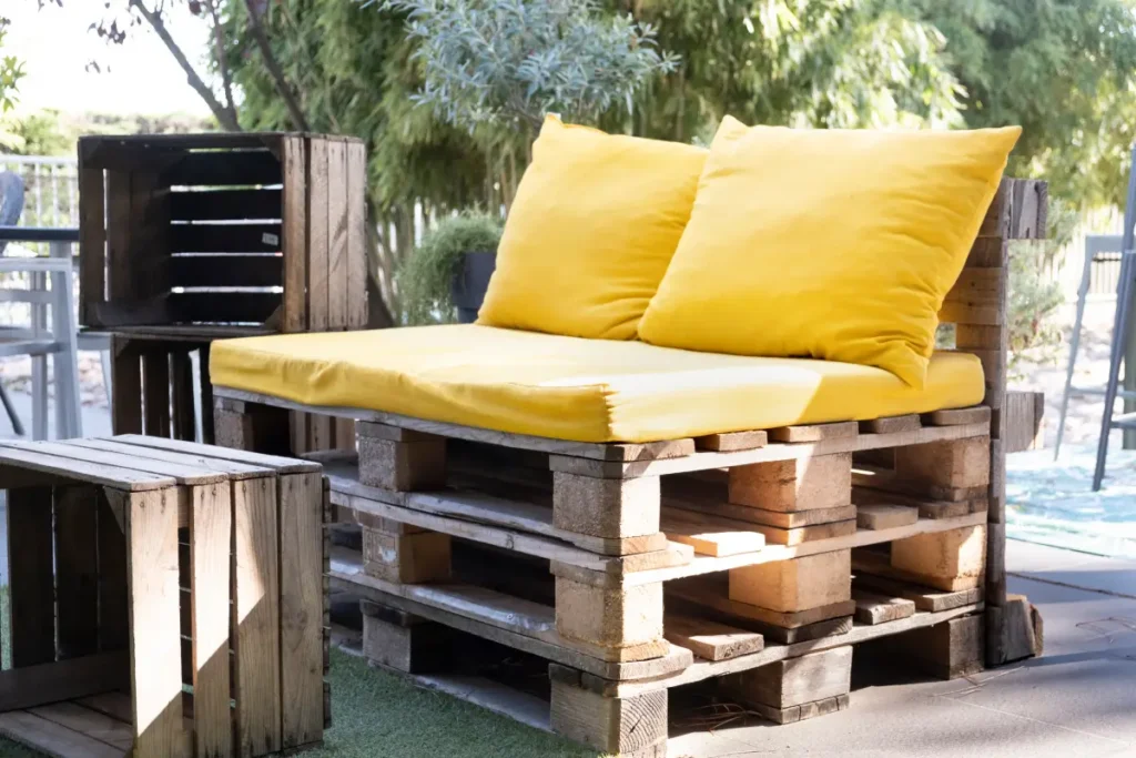 ławka z palet na tarasie z żółtymi poduszkami i stolikiem czy półkami z drewnianych pudełek
