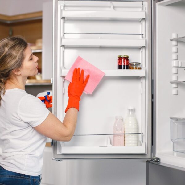 Kobieta w rękawicach myje lodówkę różową ściereczką w nowoczesnej kuchni.