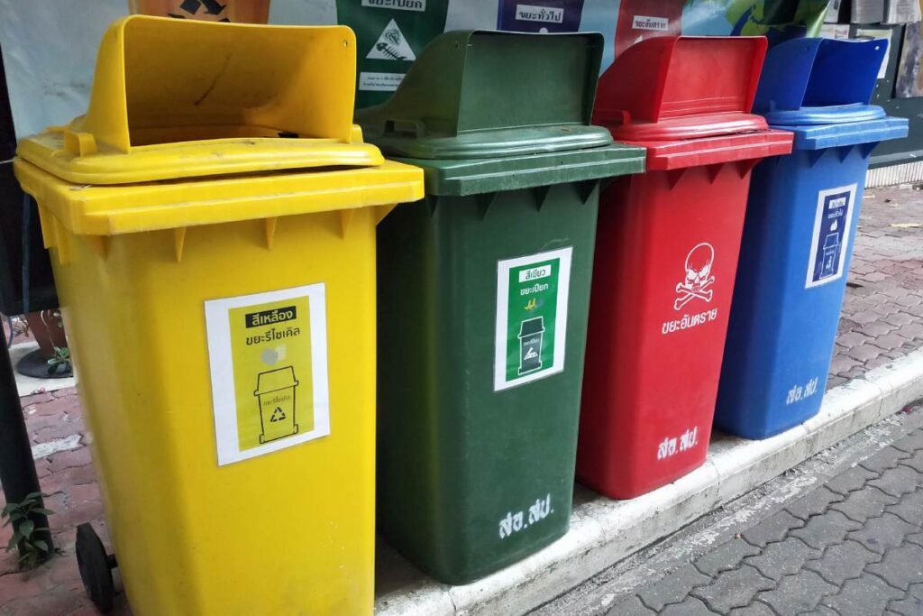 Kolorowe pojemniki na odpady: żółty, zielony, czerwony i niebieski stojące obok siebie na chodniku.