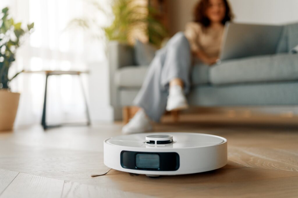Kobieta siedzi z laptopem na kanapie, obok pracuje robot sprzątający