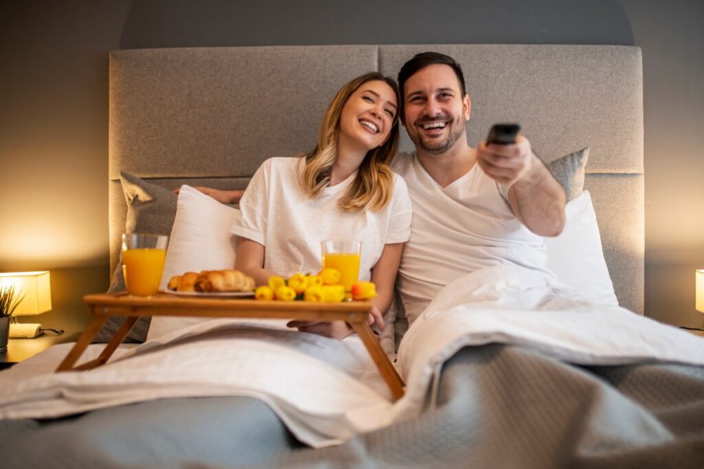 Para oglądająca telewizję w łóżku, z tacą śniadaniową i uśmiechem na twarzy