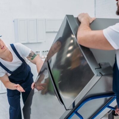 Dwóch pracowników przechyla lodówkę na wózek transportowy podczas wynoszenia jej z kuchni.